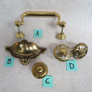 May include: A collection of brass cabinet hardware, including a handle, a cup pull, two knobs with openwork designs, and a round knob with a rope-like border. The hardware is in various shapes and sizes, all with a shiny, golden finish.