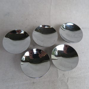 May include: Five shiny, silver-colored, concave, circular objects are arranged on a light-colored surface. The objects reflect the surrounding environment, creating a mirrored effect. The objects appear to be small dishes or decorative elements.