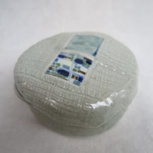 May include: A small, round, ceramic trinket box with a textured, light gray surface. The lid features a square mosaic design with blue, green, and brown tiles. The box has a slightly irregular shape and a subtle, glossy finish.