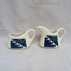 May include: A white creamer and sugar set with a blue geometric design. The creamer has a handle and a spout. The sugar bowl has a handle.