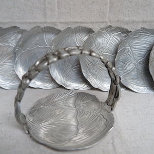 May include: Set of silver-colored metal leaf-shaped coasters with a matching handle. The coasters have a detailed leaf vein design. The handle is also silver and has a textured, twig-like appearance. A decorative item for the home.
