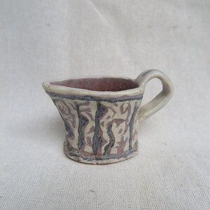 May include: A small, white ceramic pitcher with a brown handle. The pitcher has a decorative pattern of black and brown lines and shapes.