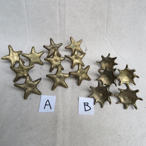 May include: A collection of brass-colored star-shaped decorative objects. Some stars have five points, while others have a spiky, multi-pointed design. The objects are arranged on a neutral-colored fabric. Two small white cards labeled "A" and "B" are present.
