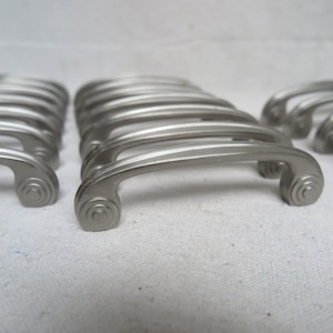 May include: A collection of brushed nickel cabinet handles. Each handle has a curved design with a scroll detail on each end. The handles are arranged in rows, suggesting they are ready for installation on furniture or cabinetry.