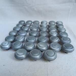 May include: A collection of thirty-six circular metal knobs arranged in a grid. Each knob features a smooth, matte gray finish with a raised central disc surrounded by a recessed ring, creating a uniform appearance across the entire set.