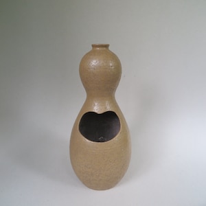 May include: A tan ceramic vase with a heart-shaped opening. The vase has a textured surface and a rounded, pear-shaped body.