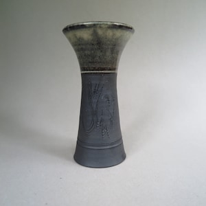 May include: A small, black and green ceramic vase with a flared top and a carved design. The vase has a dark green glaze on the top and a black glaze on the bottom.