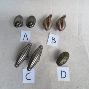 May include: A collection of antique cabinet hardware, including oval knobs, tulip-shaped pulls with copper accents, and ribbed bar pulls. The hardware is in various metallic finishes, including bronze and silver.