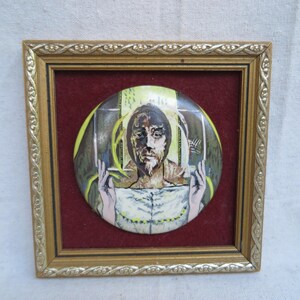 May include: A framed, circular artwork featuring a portrait of a man with a beard and raised hands. The artwork is set against a red background within a gold-colored frame. The portrait has a yellow and green background.