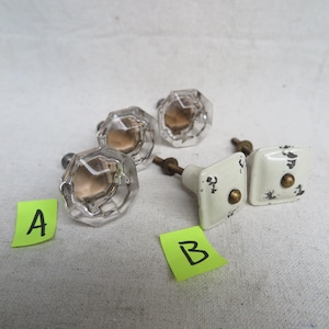 May include: A collection of vintage cabinet knobs. The knobs include clear faceted glass knobs with brass hardware and square white knobs with distressed black accents and brass hardware. The knobs are labeled A and B on yellow sticky notes.