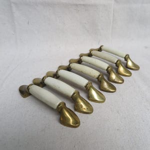 May include: A set of seven vintage cabinet pulls. Each pull features a white ceramic handle with brass-toned metal accents. The pulls are arranged in a diagonal row, showcasing their design and finish.