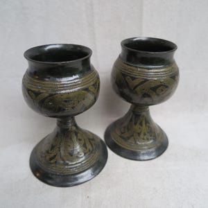 May include: Two dark green ceramic chalices with a bulbous body and a flared base. The chalices have a decorative pattern around the body and base. The chalices are handmade and have a rustic appearance.