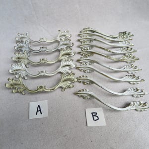 May include: A collection of ornate, vintage-style drawer pulls. The pulls are white with gold accents, featuring a curved design with floral and scroll details. Two sets are labeled A and B.