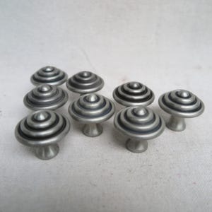 May include: Nine silver-toned cabinet knobs with a mushroom-like design. Each knob features a series of concentric rings on the top and a short, rounded stem. The knobs are arranged on a white surface.