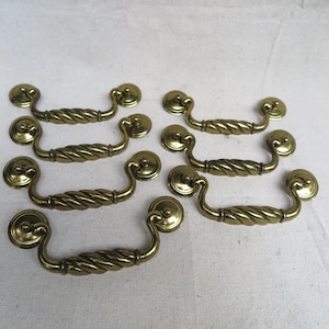 May include: A collection of six brass-toned drawer pulls. Each pull features a twisted handle design and circular mounting plates. The hardware is arranged on a white surface, showcasing their vintage aesthetic.