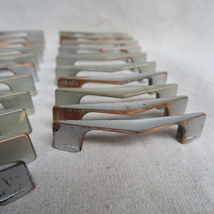 May include: A collection of multiple metal drawer pulls arranged in rows. Each pull features a central V-shaped design with a smooth, light gray finish on the top surface and exposed copper-toned metal edges showing signs of wear and patina.