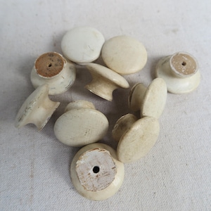 May include: A collection of ten round, off-white cabinet knobs with a smooth, slightly weathered finish. Each knob features a central hole on the flat base for mounting, with a rounded top surface. The knobs vary slightly in color and texture.