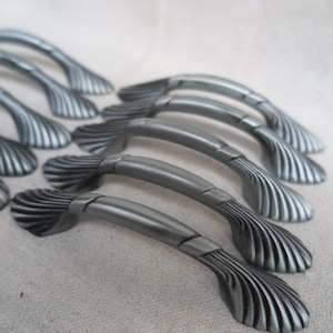 May include: A collection of gray cabinet handles with a seashell design. The handles have a curved bar and a decorative, fan-shaped end with vertical grooves. The hardware is made of metal and has a matte finish.