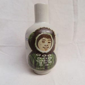 May include: A white ceramic bottle with a painted image of a person in a green parka with a fur-lined hood. The person has brown hair and a smiling face. Blue lines are painted on the side of the bottle.