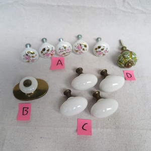 May include: A collection of decorative cabinet knobs and pulls. Includes white porcelain knobs with pink floral designs, oval white knobs with brass stems, a brass pull with a white center, and a green and gold knob.