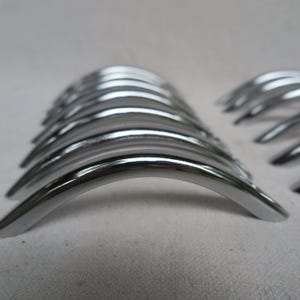 May include: A collection of shiny, curved metal objects, possibly handles or decorative hardware. The items are arranged in a row, with a polished, reflective surface. The objects are silver in color and appear to be made of metal.