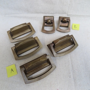 May include: A collection of antique brass drawer pulls and handles. The pulls have a rectangular design with a curved handle. Two smaller handles with a square top are also included. The hardware is aged with a vintage aesthetic.