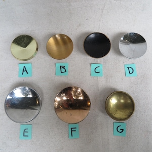 May include: A collection of seven round cabinet knobs in various metallic finishes, including gold, brass, silver, and black. Each knob is labeled with a letter from A to G on a small blue sticky note.