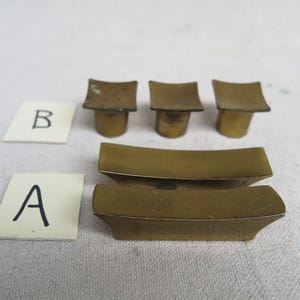 May include: An assortment of vintage brass hardware components. The image shows three small, square-topped pieces and two curved, rectangular pieces. Two white cards labeled "A" and "B" are also present. The hardware has a tarnished, aged appearance.