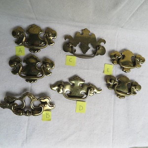 May include: A collection of six antique brass drawer pulls in various ornate designs. Each pull features a decorative handle and mounting hardware. The pulls are labeled A, B, C, D, and E on small yellow sticky notes.