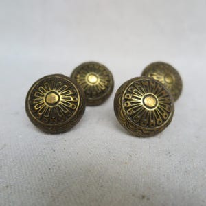 May include: Four gold-toned metal knobs with a decorative sunburst design. The knobs have a raised, textured edge.