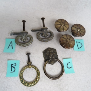 May include: A collection of antique brass cabinet hardware, including ornate pulls, ring pulls, and round knobs. The hardware features intricate designs and a vintage aesthetic. The items are labeled A, B, C, and D on blue sticky notes.