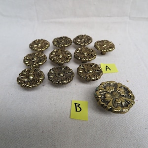 May include: A collection of antique brass floral cabinet knobs. The knobs have a detailed, embossed design with a central circular element and surrounding petal-like shapes. The knobs are arranged on a white surface.