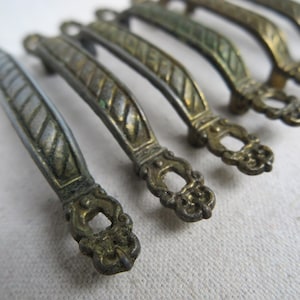 May include: A collection of antique bronze-toned drawer pulls. Each pull features a curved handle with a textured, diamond-patterned center and ornate, decorative ends. The pulls are arranged in a row, showcasing their aged appearance and intricate details.