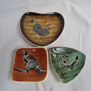 May include: Three small, decorative ceramic dishes with Australian Aboriginal art designs. The dishes are heart-shaped, square, and triangular, featuring brown, green, and tan backgrounds with black and white illustrations. The heart-shaped dish has the word "australia" written on it.