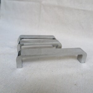 May include: Four shiny, silver-colored cabinet pulls with a rectangular, U-shaped design. The pulls are made of metal and have a smooth, reflective surface. They are arranged in a row, with one pull in the foreground and the others slightly behind.
