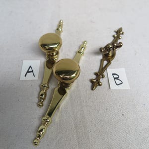 May include: Two polished brass drawer pulls labeled A, featuring round knobs on elongated backplates with tapered ends, and one ornate brass drawer pull labeled B with a decorative fleur-de-lis design. The hardware is metallic gold in color.