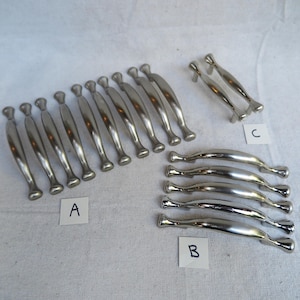 May include: A collection of silver-toned cabinet pulls in various styles. Group A features elongated pulls with a rounded top and base. Group B showcases curved pulls, and Group C presents smaller, curved pulls. All are made of a shiny, metallic material.