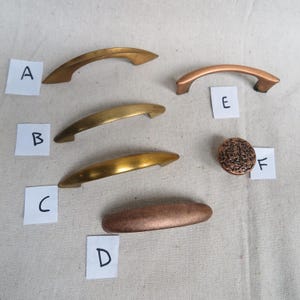 May include: Six different cabinet knobs and pulls in gold and copper finishes. The knobs and pulls are arranged in a row on a light beige fabric surface. The knobs and pulls are labeled with letters A through F.