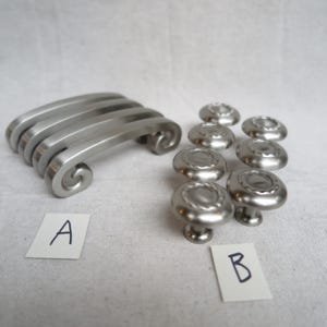 May include: A set of brushed nickel cabinet hardware. The image shows a group of handles with a scroll design and a set of round knobs. The handles are labeled "A" and the knobs are labeled "B".