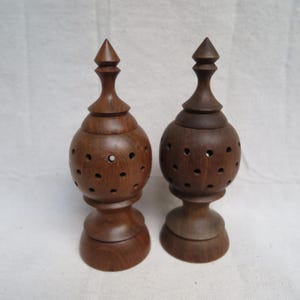 May include: Two handcrafted wooden decorative objects. Each has a bulbous body with small circular cutouts, a pedestal base, and a pointed finial. The wood has a rich, dark brown color, and the objects appear to be turned on a lathe.