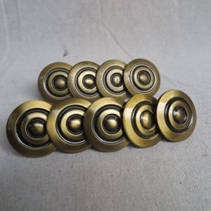 May include: A collection of eight antique brass cabinet knobs. Each knob is circular with concentric rings and a central, raised button. The knobs have a warm, aged patina, suggesting vintage hardware for furniture restoration projects.