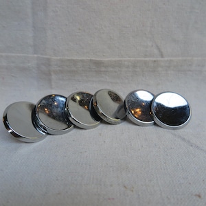 May include: A collection of six shiny, silver-colored, circular metal objects. Each object has a smooth, reflective surface, and they are arranged in a row. The objects are on a neutral-colored, textured fabric background.