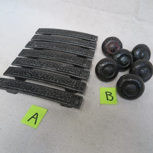 May include: A collection of dark gray cabinet hardware, including seven rectangular handles and seven round knobs. The handles have a textured surface and a curved design. The knobs have a hammered appearance.