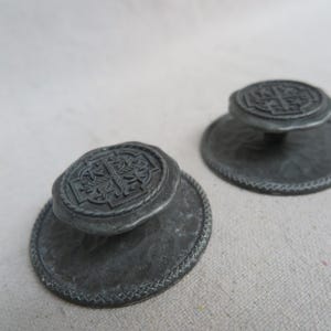 May include: Two antique metal stampers with ornate, raised designs. The stampers have a circular base with a decorative border and a detailed, oval-shaped top. The metal has a weathered, gray appearance, suggesting age and use. The design on the top appears to be a cross.