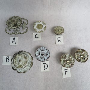 May include: A collection of six decorative metal furniture knobs labeled A through F. The knobs feature intricate floral and scrollwork patterns in various metallic finishes including gold, silver, and white enamel, each with unique circular designs.