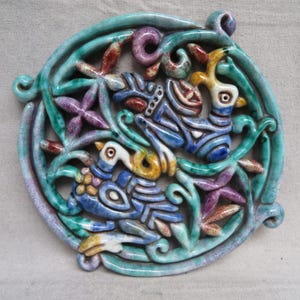 May include: A ceramic wall hanging with a circular design. The piece features stylized birds and floral elements in shades of blue, green, purple, and yellow. The artwork has a glossy finish and is likely handmade.