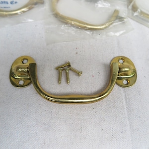 May include: A set of brass-toned drawer pulls with matching screws. The pulls have an arched handle design and oval mounting plates with screw holes. The hardware is new and ready for installation.