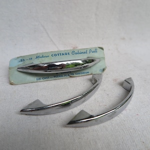May include: A set of chrome cabinet pulls with a modern design. The pulls are curved with a polished finish, and the packaging reads "Modern Cottage Cabinet Pull". The set includes two larger pulls and one smaller pull.