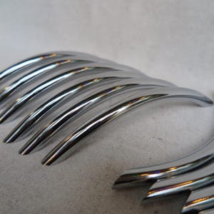 May include: Close-up of several shiny, chrome-plated metal handles. The handles have a curved, arched design, and are arranged in a row. The reflective surface of the handles creates a bright, metallic appearance. The handles are likely for furniture or cabinetry.