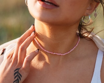 Puede incluir: Primer plano de una mujer con un top blanco sin mangas y un delicado collar de cuentas rosadas. El collar está hecho de pequeñas cuentas espaciadas uniformemente. La mujer tiene un tatuaje de una rama en su muñeca.